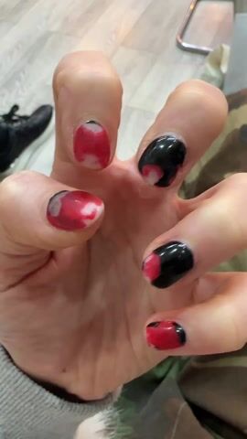 The video shows a close-up view of a hand with poorly applied red and black nail polish. The polish appears to be chipped and uneven. The hand is fair-skinned, and the fingers are slightly bent. The background includes a light-colored wooden floor, a black shoe with a clear sole, a shiny metal object, and camouflage print pants.