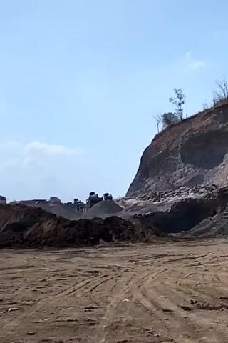 The video shows a quarry with heavy machinery. The ground is dirt with tire tracks. To the left, there are piles of dark material and several large machines, possibly trucks and crushers. In the background, a steep, rocky cliff face dominates the right side of the frame. The sky is clear and blue with a few wispy clouds. The overall impression is of an active industrial site in a natural landscape.