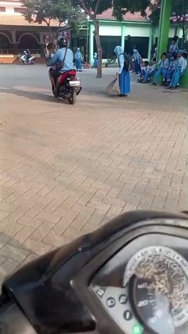 The video shows an outdoor scene, possibly a school courtyard, with several individuals present. In the foreground, the dashboard of a motorcycle is visible, suggesting the footage is being recorded from the perspective of someone on the motorcycle. The background features buildings, trees, and a paved area. Several people, mostly young individuals in what appear to be school uniforms, are scattered across the scene. Some are walking, others are standing, and a few are seated. One individual, dressed in a blue hijab and uniform, is seen sweeping the ground with a broom. Another person is riding a motorcycle in the left portion of the frame. The overall atmosphere seems to be that of a school environment during what appears to be daytime, with sunlight casting shadows.