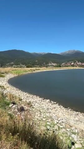 A slow panning shot reveals a tranquil lake bordered by a rocky shore and sparse vegetation. In the background, a verdant hillside is dotted with trees, and a small town with red-tiled roofs is nestled among them. The scene is completed by a range of distant, majestic mountains under a clear, bright blue sky.