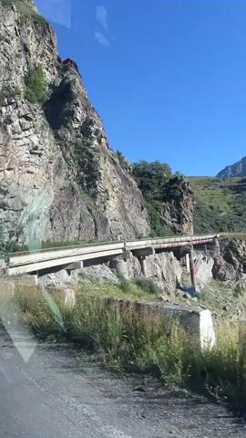 The video shows a car driving along a narrow mountain road. The road is carved into the side of a steep cliff, with a sheer drop on one side. The cliff face is rocky and uneven, with some vegetation growing in the crevices. The road is made of asphalt and has a low concrete barrier on the edge. There is a bridge-like structure crossing a gap in the cliff, with metal supports and a concrete deck. The sky is clear blue and the sun is shining. The mountains in the distance are covered in green vegetation. The car appears to be moving slowly, navigating the tight turns of the road. The overall impression is one of a scenic but potentially dangerous drive.