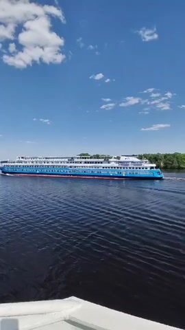 A cruise ship sails down a body of water on a clear day. The sky is blue with puffy white clouds. The water is dark and rippled. The cruise ship is blue and white with several decks. Trees can be seen on the right side of the frame. The bottom of the frame shows the side of another boat, from which the video appears to have been taken.