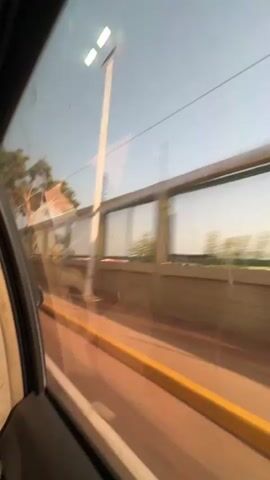 The video shows a view from the backseat of a moving vehicle, likely a car or minivan. A person's head is visible in the foreground, resting against a beige headrest with a small television screen. The camera focuses on the window. Outside, a sound barrier runs alongside the road. The barrier is constructed of horizontal panels with alternating wooden slats and semi-transparent, light-colored plastic sheets. A yellow line marks the edge of the road. The sky is clear and light blue, indicating daytime. There are trees and other buildings visible in the distance. The interior of the vehicle is beige and gray.