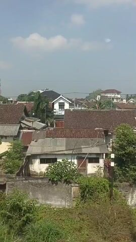 The video shows a view of a densely populated residential area with many houses. The houses have tiled roofs, some of which appear to be in disrepair. There are trees and vegetation scattered throughout the scene. The sky is visible, with some clouds. The overall impression is of a rural or semi-urban environment.