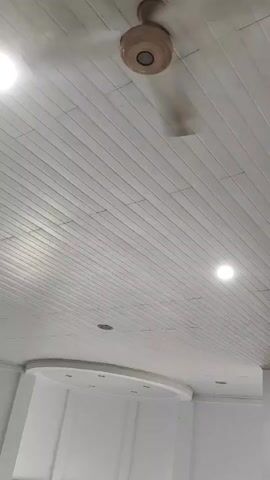 This is a very short video, lasting only three seconds, showing a ceiling with a white paneling and a fan in the center. The fan appears to be made of wood and metal and is spinning, indicating movement. There are also two circular lights embedded in the ceiling on the right side of the frame, and a circular recessed area with more lights on the left side. The overall environment appears to be an indoor space with a clean, modern aesthetic.