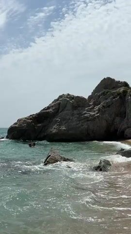 The video shows a beach scene with a large rock formation on the left and a sandy shore on the right. The water is clear and turquoise, with small waves breaking on the shore. There are a few people swimming in the water near the rocks. In the background, there is a small stone structure built into the side of the cliff. The sky is blue with scattered white clouds. The video appears to be taken on a sunny day.