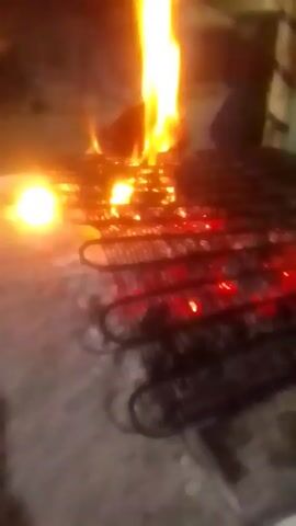 The video shows a metal rack or grid on fire. The flames are bright orange and yellow, with some areas glowing red underneath the metal. The rack is sitting on a concrete surface, and the background is dark, suggesting it is nighttime. There are some blurry lights visible in the distance, but they are out of focus. There are no people visible in the frame. The focus is mainly on the fire and the metal rack.
