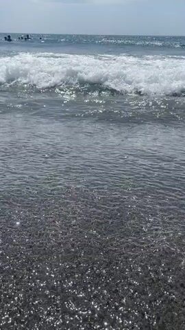 A video of a beach on a sunny day. The camera is positioned low to the ground, looking out towards the ocean. In the foreground, dark sand sparkles with tiny white flecks, possibly mica or shells. A wave breaks gently on the shore, its white foam dissolving into the dark, wet sand. In the middle ground, the water is a shimmering blue-gray, with several people visible in the distance, appearing to be swimming or playing in the ocean. The sky above is a pale blue with a few wispy white clouds. The video captures the tranquil beauty of a coastal environment.