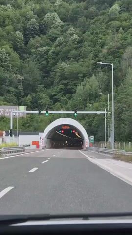 The video shows a first-person perspective of driving towards and entering a tunnel. Green traffic lights are visible overhead, indicating that it is safe to proceed. The tunnel entrance is illuminated, and speed limit signs displaying "80" are visible on either side of the tunnel. The tunnel walls are lined with lights and what appear to be warning signs or markers. The surrounding environment outside the tunnel entrance is mountainous and covered in dense green trees. A sign in Bulgarian language is visible on the left side of the road, near the tunnel entrance.