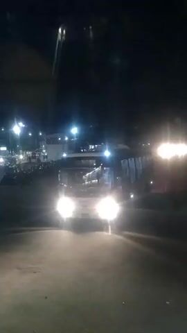 The video depicts a night scene at what appears to be a bus terminal or parking area. A brightly lit bus pulls into the frame, its headlights shining forward. The camera angle is from a slightly elevated position. As the bus turns to its right, it reveals three individuals on a motorcycle, also brightly lit with its headlights. In the background, there are more lights suggesting other parked vehicles and possibly a building. The overall atmosphere is dark due to the night, but illuminated by various vehicle and street lights.