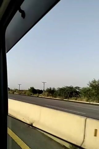 The video shows a view from inside a vehicle looking out the window at a highway. The highway has a concrete barrier separating the lanes. There are utility poles and sparse vegetation along the side of the road. Several cars are seen driving on the highway. The sky is clear and blue. The video is short, lasting only a few seconds, and the camera appears to be moving.