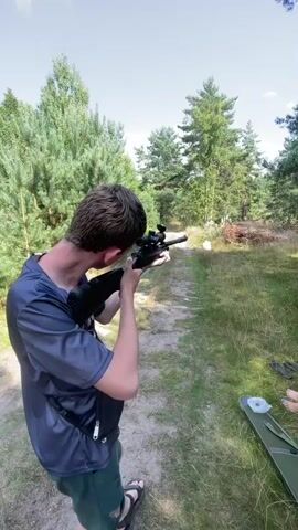 A person is aiming a rifle down a dirt path in a wooded area. The person is wearing a dark blue t-shirt and green shorts. There are trees and grass on either side of the path. The sky is visible and appears to be partly cloudy.