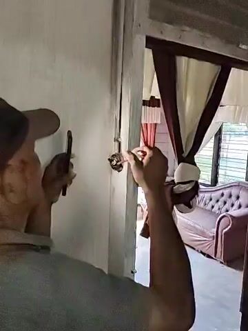 A man wearing a cap and a gray shirt is standing in front of a wall. He is using a small tool to work on a small, circular object attached to the wall. In his other hand, he is holding a phone.  Through the doorway behind him, a woman wearing a white head covering and maroon shawl is visible next to a tufted, rose-colored couch. Light comes through the window behind her. The wall is painted white, and the doorway is made of raw, unfinished wood.