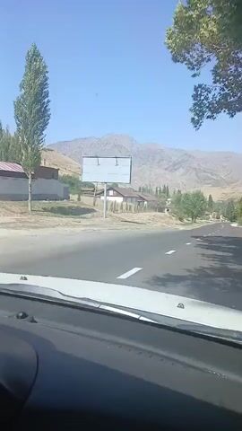The video shows a view from inside a car driving on a paved road. On the left side of the road, there is a billboard and some houses. On the right side of the road, there are trees and a few houses. In the background, there are mountains. The sky is clear and blue. The video captures the car driving forward, with the scenery changing as the car moves. The camera seems to be mounted on the dashboard of the car. There are no people visible in the video. The overall environment appears to be rural or semi-rural, with a natural landscape dominating the scene. The sunlight suggests it is daytime.