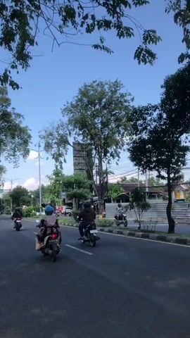 The video shows a street scene with several people riding motorcycles. The road is paved and has a yellow line marking the edge. Trees line the side of the road, and there are buildings in the background. The sky is blue and clear. The motorcycles are moving in the same direction, away from the camera. One motorcycle in the foreground has two people riding on it, a driver and a passenger. The driver is wearing a helmet and a long-sleeved shirt. The passenger is wearing a headscarf and a patterned shirt. Another motorcycle in the middle ground has a single rider wearing a helmet and a dark jacket. Several other motorcycles are visible further down the road, also moving away from the camera. The overall impression is a busy street on a clear day.