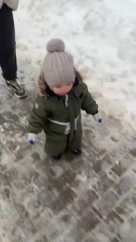 A man and a young child are walking outside on a snowy day. The child, wearing a green snowsuit and gray hat, toddles along the sidewalk. The man, wearing a beige puffer jacket and dark pants, walks beside the child with his hands in his pockets. The sidewalk is icy. The background shows snow-covered ground and a fence.