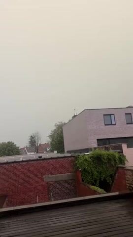The video is a short, shaky clip of an overcast sky above a residential area. The sky appears to be a uniform light gray, suggesting cloudy or stormy weather. In the middle ground, there are several rooftops, with one modern-looking building made of light-colored brick on the right, featuring black-framed windows. To the left of this building, there is a red brick building with a rooftop covered in greenery. In the foreground, a wooden surface, possibly a deck or roof, is visible. Trees can be seen between the buildings on the left. The overall impression is of a gloomy, possibly rainy day.
