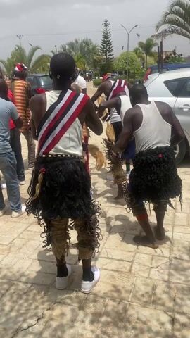 In an outdoor setting with palm trees and vehicles nearby, a group of men engage in what appears to be a traditional dance. The central figures are adorned in distinctive attire: white tank tops, short skirts made of dark fringed material, and leg decorations consisting of woven fibers and metallic elements. One dancer has a sash with diagonal red, white, and black stripes. Some men clap their hands and follow the dancers' movements. The surroundings include a paved area and partial views of a building in the background. The overall impression is a cultural performance or celebration.