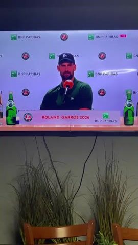 The video shows a person sitting at a table and speaking into a microphone. The person is wearing a green shirt and a dark blue baseball cap. There are two bottles of Perrier sparkling water on the table, as well as a bottle of wine. Behind the table is a backdrop with the BNP Paribas logo and the words "Roland Garros 2026." Below the table are three chairs and tall grass-like plants. The person on screen appears to be giving a press conference.