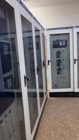 The video shows a room containing electrical control panels. The panels are white with glass doors and have various buttons and displays inside. A desk with a computer, printer, and other equipment sits in the room. There are also a few other cabinets and a fire extinguisher. The room has tiled floors and white walls. The camera pans slowly from left to right, showing the extent of the electrical equipment in the room. There is no one visible in the room.