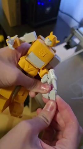 In a close-up shot, two hands are seen holding a yellow and white robot toy, resembling Bumblebee from Transformers. The robot is positioned vertically, with its legs bent and its arms extended. One hand is supporting the robot's body, while the other hand is manipulating a small, gray accessory, possibly a weapon, near the robot's right hand. The accessory appears to be a gun-like object. The background is blurred but suggests an indoor setting with a desk, a computer tower with blue LED lights, and a wooden surface. The lighting is moderate, casting some shadows. The focus is on the action of attaching or detaching the accessory to the robot toy.