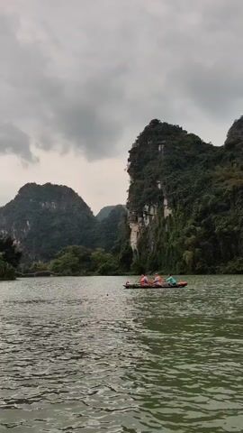 A distant shot shows a body of water on an overcast day with several rocky, tree-covered hills in the background. In the middle of the water, a long boat with several people wearing life vests can be seen. Another boat is seen passing by in the foreground. The water surface is rippled.
