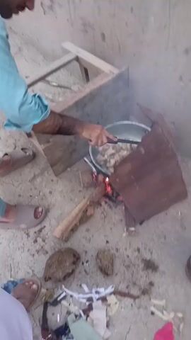 The video shows an outdoor scene in Kakki, Pakistan, where someone is cooking food over an open fire. The setting appears to be rustic, with a dirt ground and a simple setup for cooking. A metal pot is placed over a fire made with wood. The person cooking is using a utensil to stir the food in the pot.

Visible subjects include parts of people's feet wearing sandals, suggesting they are standing around the cooking area. Objects in the scene include the cooking pot, wood for the fire, some debris on the ground, and a wooden structure in the background that might be furniture or part of a shelter.

The activity is clearly cooking, likely a meal being prepared outdoors. The time of day is not explicitly indicated, but the lighting suggests it could be daytime. There are no clear indicators of weather conditions. The location cues, such as the dirt ground and the type of cooking setup, point to a rural or less developed area, consistent with the provided location context of Kakki, Pakistan.

Notable details include the presence of smoke rising from the fire, indicating that cooking is in progress. The food in the pot appears to be pieces of meat or vegetables. The overall impression is one of simple, traditional outdoor cooking. There is no visible text in the video. The emotions are neutral, focusing on the act of cooking.