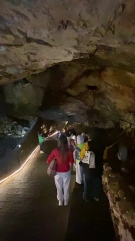 A group of people are walking through a cave. The cave walls and ceiling are made of rough rock. A pathway runs through the cave, and a strip of light lines the edge of the pathway. The people are walking away from the camera. They are wearing various types of clothing.  Some of them are carrying bags. One person is wearing a green jacket, and another is wearing a red jacket and white pants. The lighting in the cave is dim.