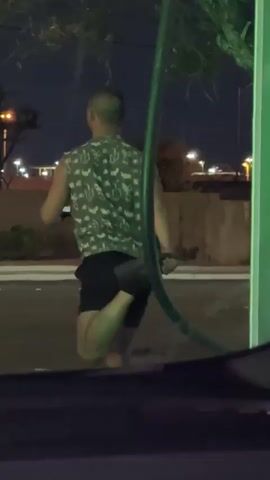The video shows a man from behind, illuminated by nearby lights in the dark. The man has a short haircut and is wearing a sleeveless shirt with a cactus pattern and dark shorts. He is barefoot and appears to be warming up or stretching his leg, holding his foot behind him. He is on a paved surface. A green hose or pipe is in the foreground, partially obstructing the view. Behind the man is a low wall and some foliage. In the distance, lights from buildings or street lamps are visible. There are also moving headlights in the background.