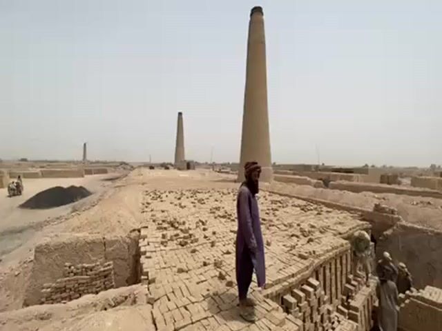 The video shows a brickyard. A man with a beard and wearing a head covering and a long purple shirt is standing on a pile of bricks. He looks to the right. There are other men on the right who are working with bricks. There are three tall chimney towers in the background. All around are stacks of bricks. The scene is dry and dusty.