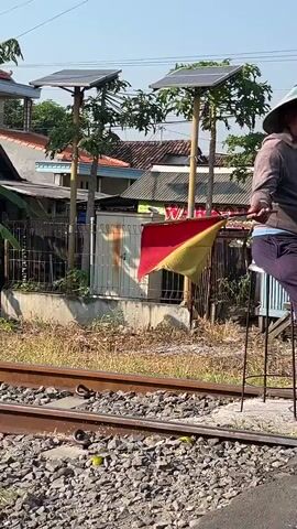 An elderly man wearing a conical straw hat and a gray hooded sweatshirt sits on a black stool next to a set of train tracks. He is looking to the right of the frame and holding a yellow object in his right hand. His left hand is not visible. He is wearing dark pants and has a blue waistband visible. Behind him, there is a fence, some trees, and houses with solar panels on their roofs. The ground is covered with gravel and dry grass. The lighting suggests it is daytime.