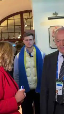 The video shows a man standing indoors wearing a blue and yellow scarf, a white puffer vest over a light blue shirt. He is surrounded by other people, including a woman in a red jacket and a man in a dark suit. The background shows a room with a window and a wooden sign. The man in the scarf appears to be listening to someone out of frame. He has a serious expression.