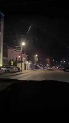 The video shows a night-time view from inside a car driving down a city street. The road is lined with parked cars on both sides. Streetlights illuminate the road and the buildings.  There are a few cars driving in the opposite direction.  The windshield has some streaks or dirt on it, possibly from rain or moisture. The dashboard is visible at the bottom of the frame.  Music is playing in the background.