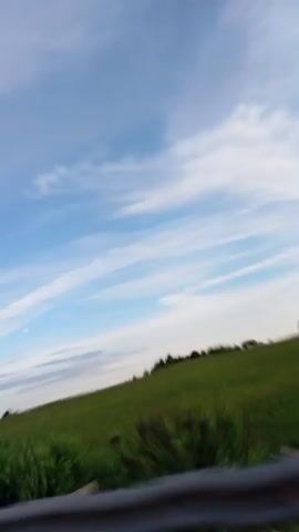 The video shows a view of a grassy field and sky from a moving vehicle. A metal guardrail is visible in the foreground. The sky is blue with scattered white clouds. The field is green and appears to be on a hill. Trees are visible in the distance. The video is shaky, suggesting it was recorded from a car or other moving vehicle.