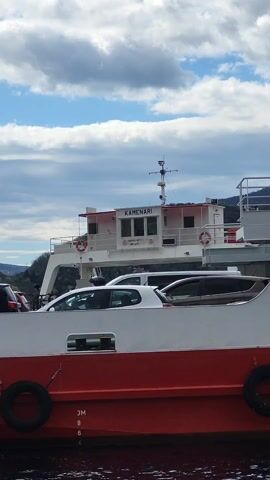 The video captures a static, medium shot of a car ferry named "KAMENARI." The ferry is painted in red and white, with cars loaded on its deck. The focus is primarily on the ferry's structure, with the name "KAMENARI" prominently displayed on the cabin. The sky above is partly cloudy, with patches of blue visible. The background includes a blurred view of a shoreline with trees. The overall scene appears to be taken during daytime.