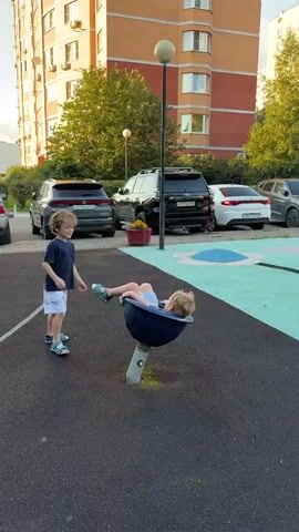 Two young boys are playing on a playground. One boy, wearing a dark blue t-shirt and white shorts, stands to the left of a spinning toy. The other boy, wearing a blue t-shirt and blue shorts, is seated in the spinning toy. The boy on the left is pushing the spinning toy, causing the boy on the right to spin. The playground has a dark asphalt surface and a turquoise and pink play area in the background. Several cars are parked behind the playground, and a tall, multi-story brick building is visible in the background. A streetlamp stands near the spinning toy.