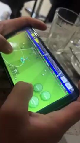 A close-up shot shows a person playing a soccer video game on a smartphone. The screen displays a green soccer field with players in action. The person's fingers manipulate virtual controls on the screen. The phone is held close, obscuring the background, but hints of a table, glassware, and another person are visible.