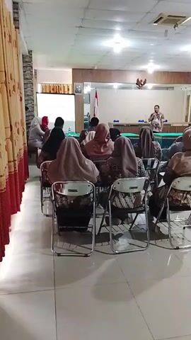 A man in a patterned shirt stands at a podium speaking to an audience seated in chairs. The audience consists of men and women, many of whom are wearing headscarves. The room is well-lit with fluorescent lights and appears to be a conference room or classroom. There is a projector screen behind the speaker and Indonesian flags visible on the wall. The chairs are arranged in rows facing the front of the room. The overall atmosphere suggests a presentation or lecture is taking place.