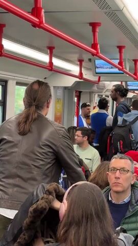 The interior of a train is shown in a slightly high-angle shot. The train is crowded with standing and sitting passengers. Red bars line the ceiling, which is lit by fluorescent lights. Some of the passengers are looking at their phones or devices, while others are conversing. A television monitor is above the heads of the passengers. The train windows are visible on the left side.