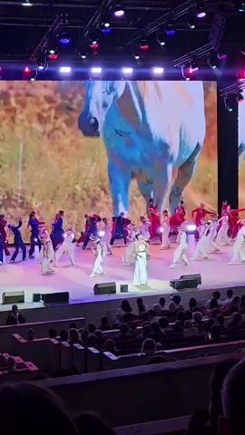 A live performance is taking place on a stage in front of an audience. The stage features a large LED screen displaying various scenes, including a close-up of a white horse's head and a scene with people on horseback by a body of water. On the stage, there are many performers in white and red costumes, engaged in choreographed dance movements. In the center of the stage, a woman in a white dress stands alone, seemingly singing or speaking. The audience is seated in a darkened theater, with rows of chairs visible. Stage lighting illuminates the performers and the screen.