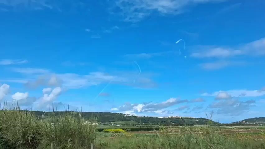 The video appears to be taken from inside a moving vehicle. The view is through the windshield, showing a bright blue sky with scattered white clouds. In the foreground, there's green vegetation and tall grasses. Further in the distance, a slightly elevated land area is visible, with some buildings or structures dotted across it. A guardrail is seen alongside the road.  The overall scene suggests a rural landscape on a clear, sunny day.