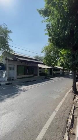 This is a vertical video filmed on a sunny day in Nganjuk, Indonesia. The scene is a street with a building on the left and trees on the right. A paved road runs through the center of the frame, with a sidewalk on the right. The building appears to be a shop or restaurant, with a corrugated metal roof and a light-colored facade. There is a white gate to the left of the building. The trees on the right are full and green, casting shadows on the road.

The activity taking place is a person riding a motorcycle down the street. The rider is wearing a helmet. A white car is also visible further down the road. The sky is clear and blue, indicating good weather. The sunlight suggests it is likely mid-morning or early afternoon. The overall mood of the video is peaceful and ordinary, capturing a typical moment in a roadside setting. No text is clearly legible within the video.