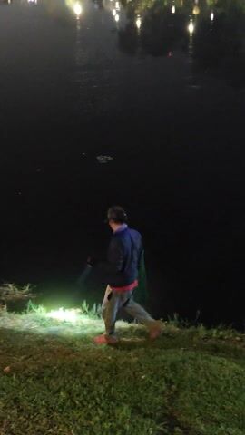 At night, a person walks near a body of water. The person is wearing a dark jacket, light pants, and red shoes. They carry a fishing net in one hand and a flashlight in the other, which shines on the grass at their feet. The water is dark with reflections of lights visible in the distance. The person is walking along the grassy edge of the water.