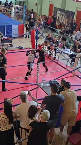 The video shows a kids’ kickboxing match in a ring surrounded by spectators. Two young fighters wearing protective headgear and shin guards are actively engaged in combat. The ring has a red floor, and blue and white padded corners, secured by metal poles and red and white ropes. Referees in black uniforms monitor the match closely. Spectators, some holding up their phones to record, are seated and standing around the ring, separated by a wire fence. The setting appears to be an indoor gymnasium or sports hall, with flags and banners visible in the background. There's a water bottle and other objects on the floor near the ring.