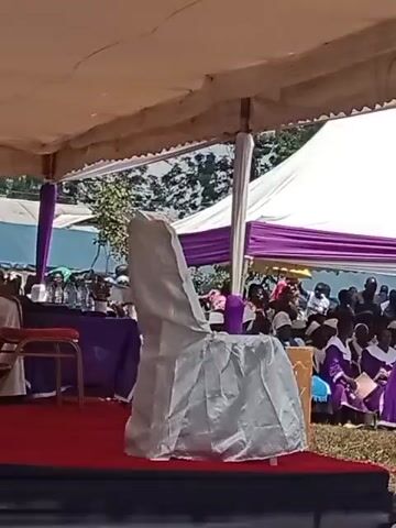 The video shows a man wearing a long white robe and white clerical collar walking on a stage. He is walking from a red chair to a table covered with a purple cloth and bottled water.  The stage is covered with a red rug and is beneath a large off-white tent. A chair covered in silver cloth is nearby. In the background are many people sitting under a second white tent with purple accents. Trees and natural vegetation are visible behind the tents.