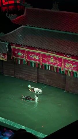 The video shows a Vietnamese water puppet show. Two puppet figures, one of a man on a white ox and the other of a man on a black duck, are in the water. The stage is decorated with traditional Vietnamese architecture and designs. Live musicians can be seen to the side of the stage.