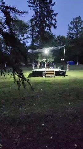 The video begins with a wide shot of an outdoor stage set up in a grassy area with trees in the background. The sky is dark, indicating it is either dusk or night, and the stage is lit by spotlights. A few people are visible on and around the stage, including one person who appears to be speaking into a microphone. As the video progresses, the camera pans slightly to the right, revealing an audience seated on the grass in front of the stage. The audience members are mostly adult, and appear to be watching the performance. Several speakers are positioned on either side of the stage. The overall atmosphere suggests an outdoor concert or event.