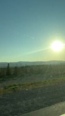 The video shows a view from inside a vehicle looking out a window at a bright sun shining in a clear blue sky. The sun is slightly off-center to the right, and its rays are creating a lens flare effect on the window. In the distance, there are rolling hills or mountains. The foreground shows a landscape of green grass and dirt road. The window appears to be dirty or scratched, with visible marks and smudges. The overall impression is that of a sunny day during a drive, possibly at sunrise or sunset given the angle of the sun.