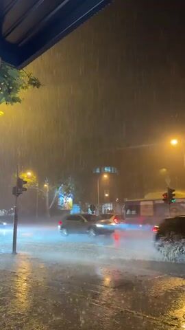 A night-time scene of a heavy rainstorm at an urban intersection. Water covers the road, creating reflections of streetlights and vehicle headlights. Several cars are driving through the flooded intersection, kicking up spray. A traffic light is visible on the left, and a bus is passing in the background on the right. The atmosphere is dark and obscured by the rain.