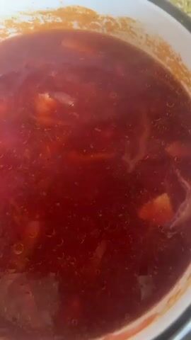A close-up overhead shot shows a pot of simmering red soup, possibly borscht. Steam rises from the liquid, which is a deep red color with small bubbles on the surface. A spoon is used to stir the soup, lifting pieces of cabbage and other vegetables. The inside of the pot appears to be white enamel with some browning on the rim. The pot is sitting on a black stovetop.